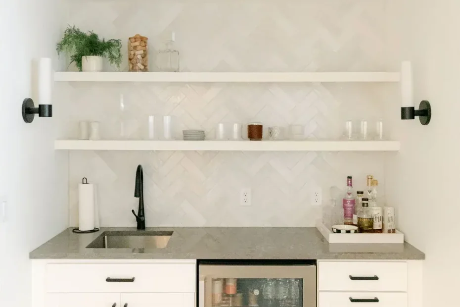 White wet bar with grey counter, herringbone tile backsplash, floating shelves and glass-front beverage fridge