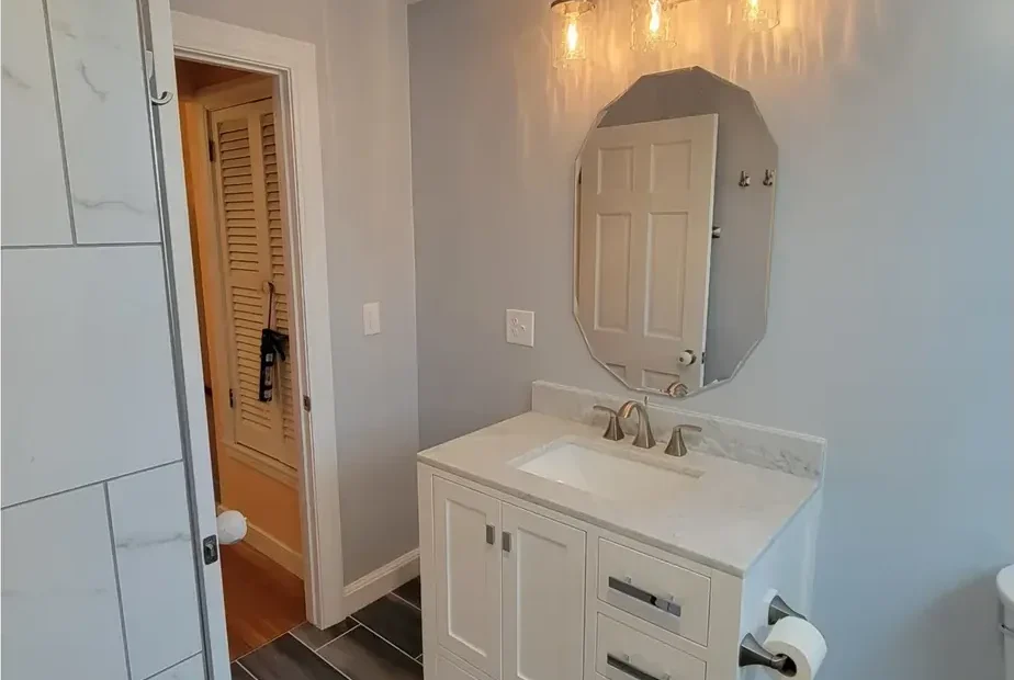 Finished bathroom with white vanity, stone-look top, octagonal mirror, sconce and grey wood-look tile floor