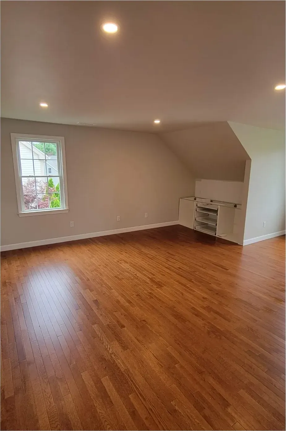 Empty sloped-ceiling room with hardwood floor, painted walls, recessed lights and open built-in shelving under the eave