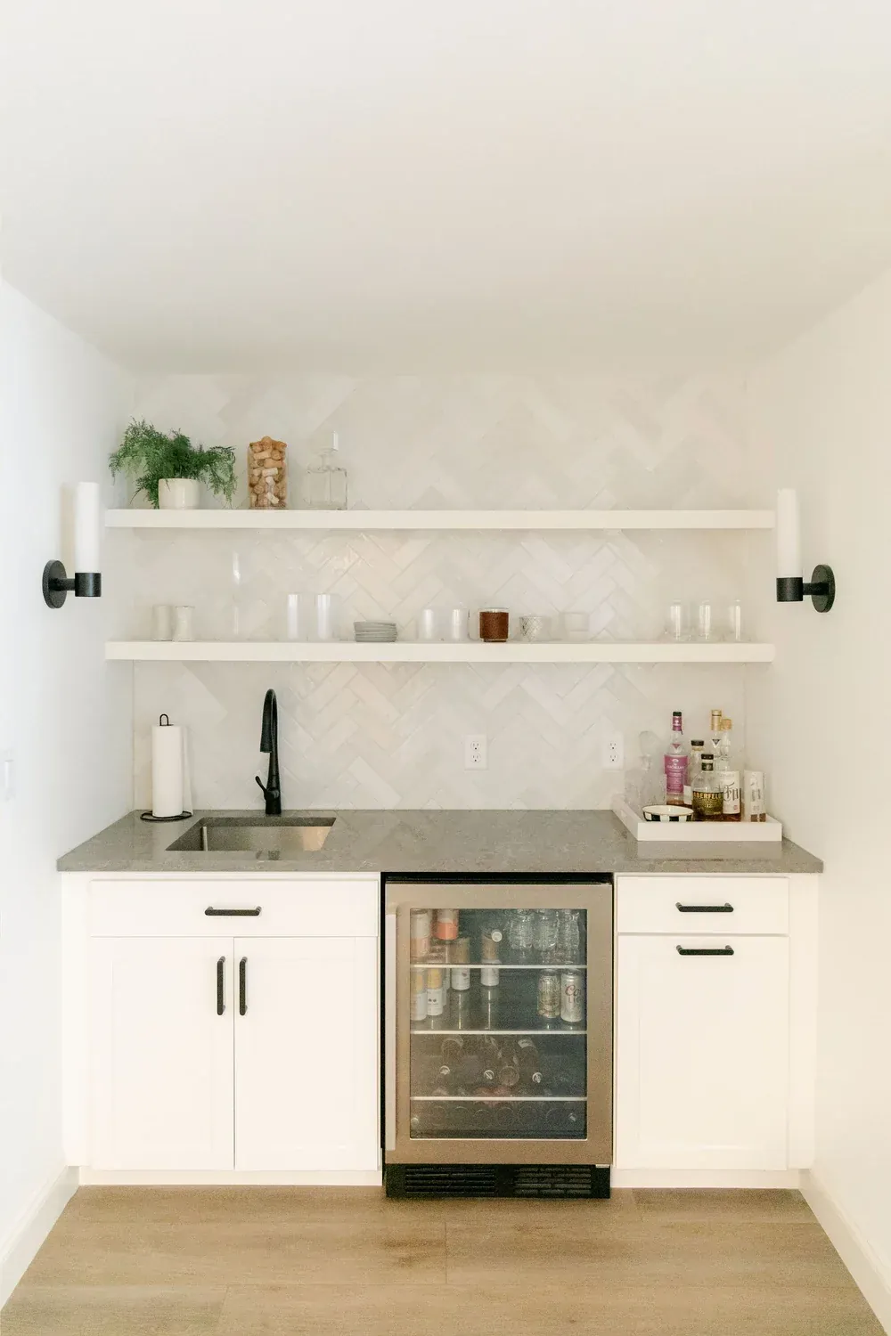 White wet bar with grey counter, herringbone tile backsplash, floating shelves and glass-front beverage fridge