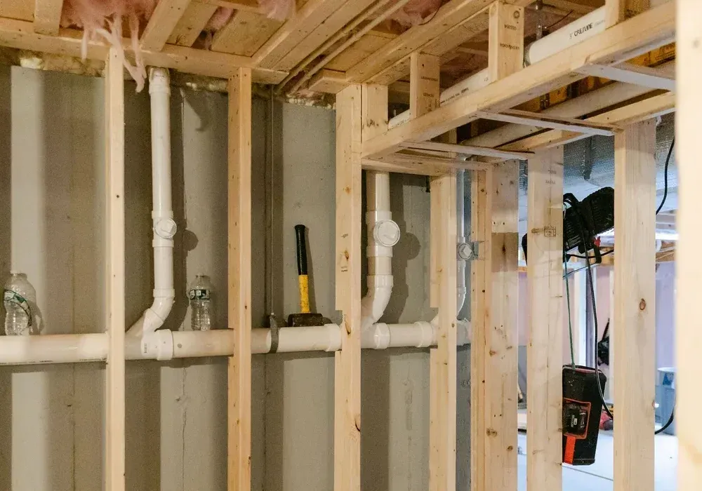 Basement framing against concrete walls with white PVC plumbing lines, sledgehammer and water bottles on studs