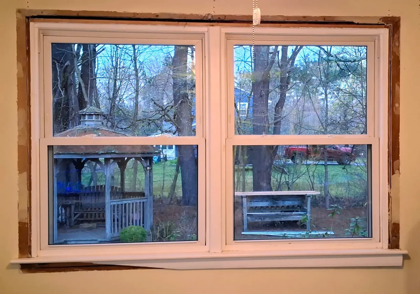 New twin double-hung window set in a tan wall with unfinished edges, wooded yard and gazebo beyond