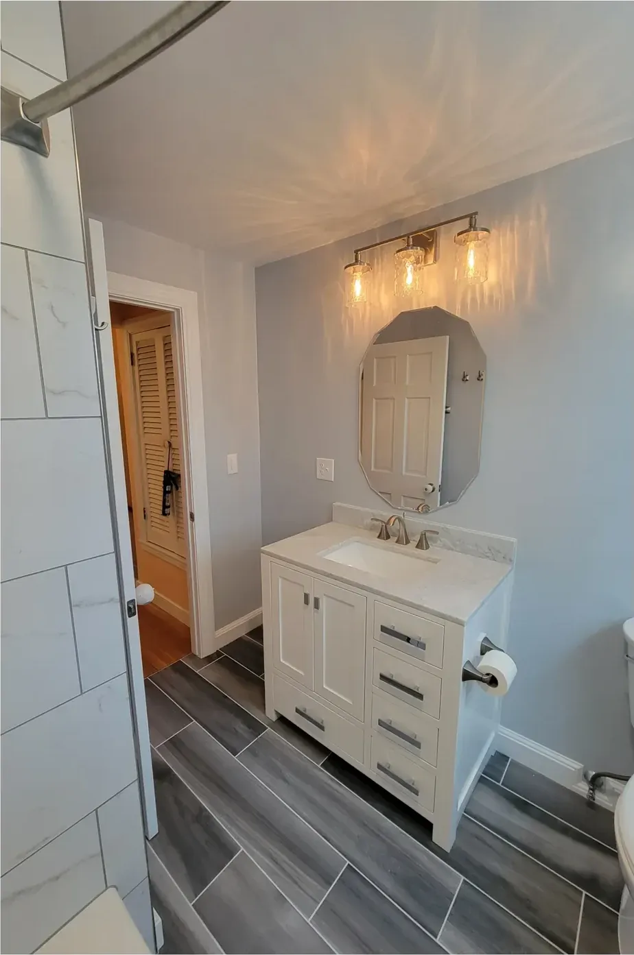 Finished bathroom with white vanity, stone-look top, octagonal mirror, sconce and grey wood-look tile floor