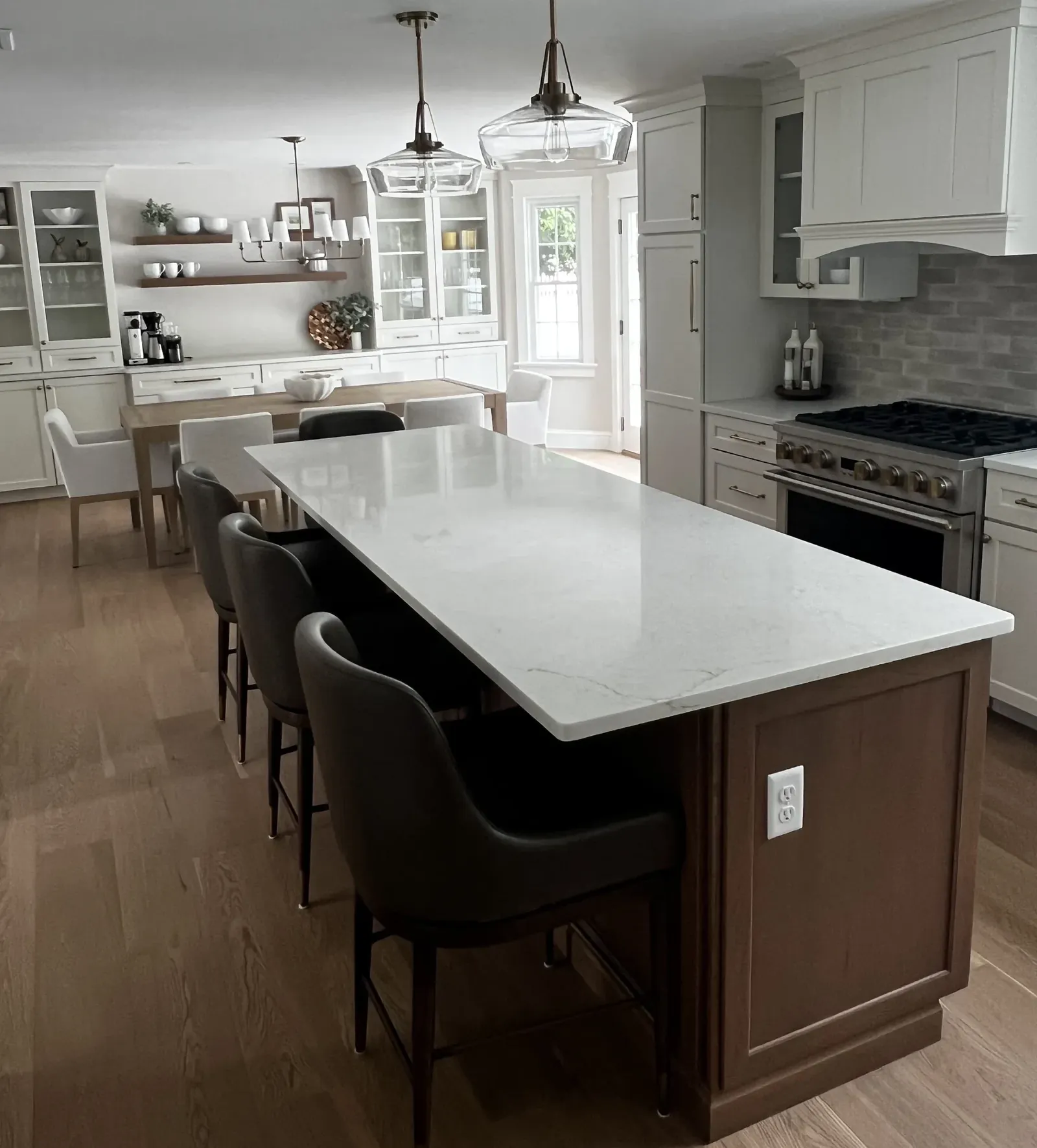 White kitchen with long veined stone island, dark stools, glass pendants, gas range, and dining table beyond