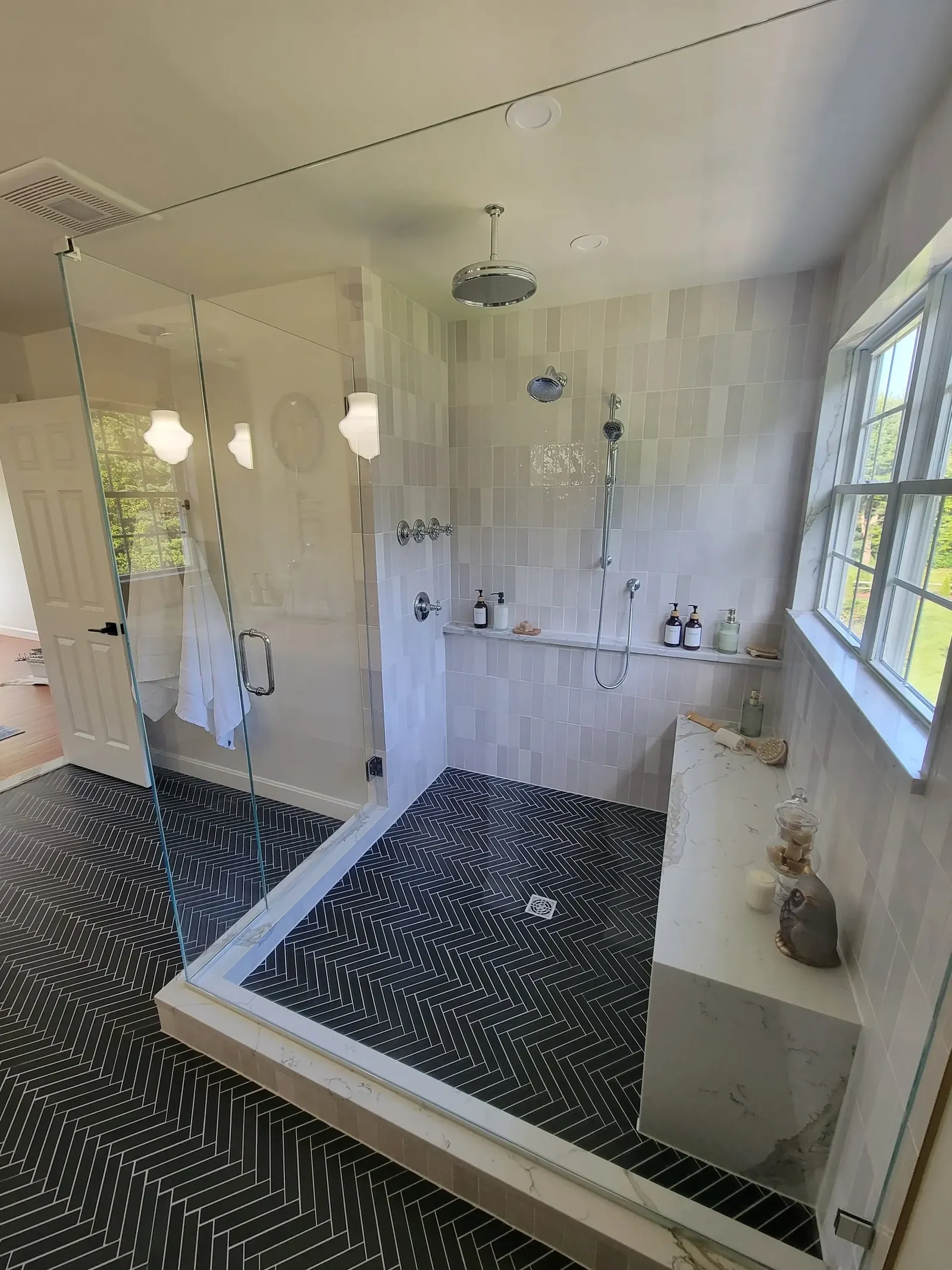 Walk-in glass shower with white stacked tile walls, black herringbone floor and chrome rain showerhead